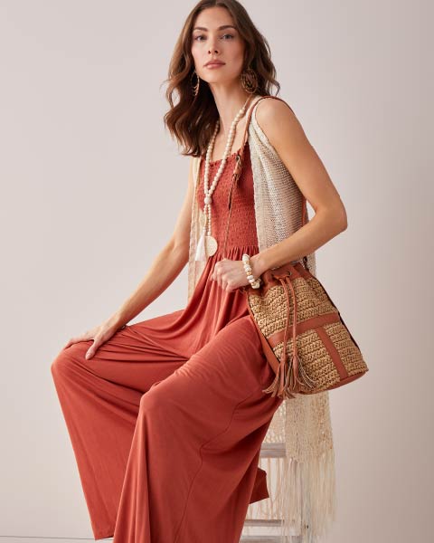 Seated model wearing amber color romper and open knit sleeveless sweater and holding satchel bag